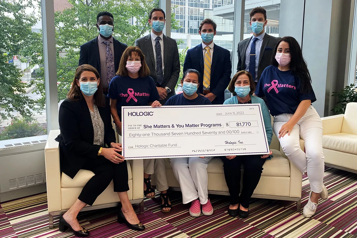 Foto de grupo de indivíduos mascarados a segurar um grande cheque promocional dos programas «She Matters & You Matter»